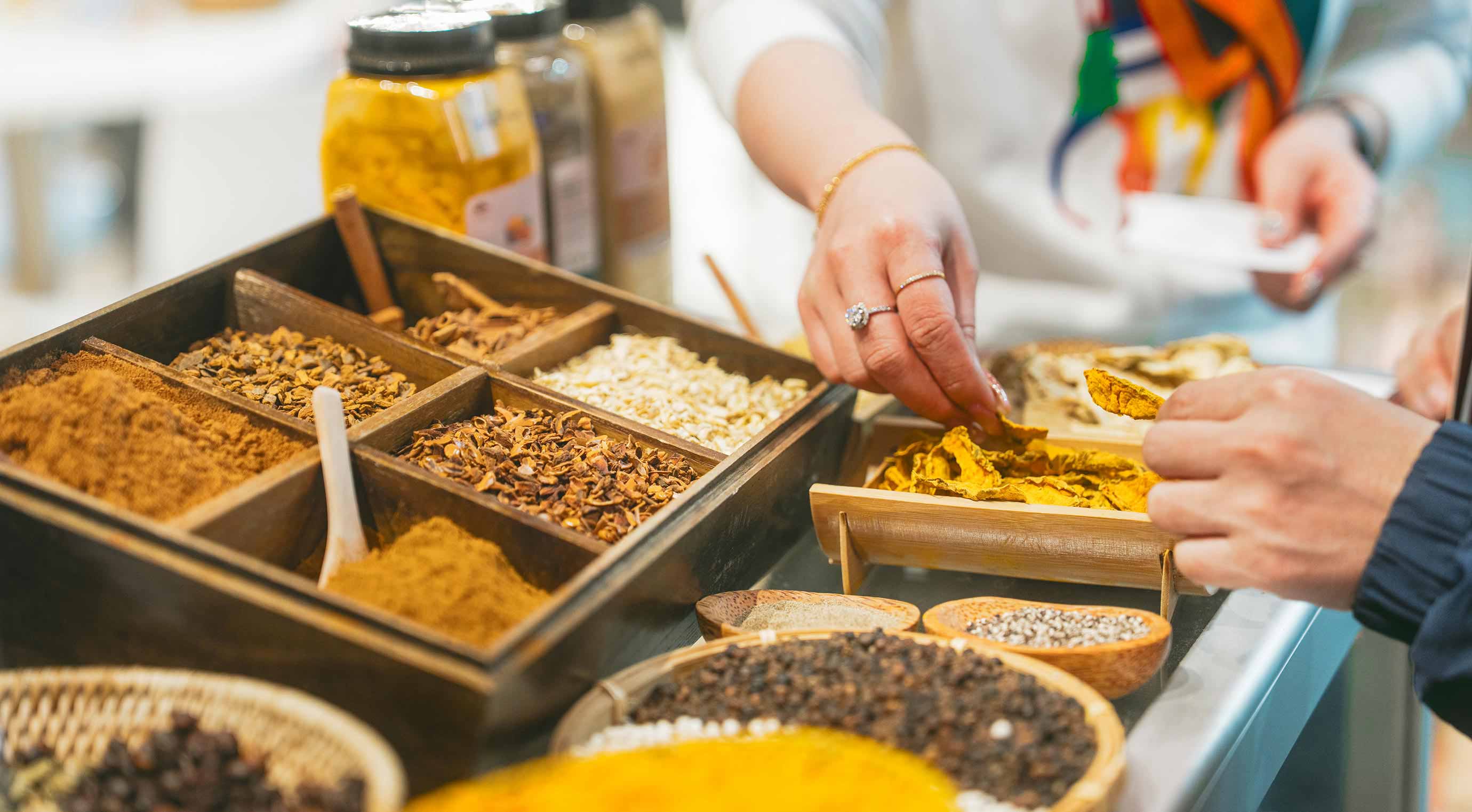 Nahaufnahme einer Hand, die nach einer Trockenfrucht greift. Zu sehen sind weitere Trockenfrüchte und Gewürze.