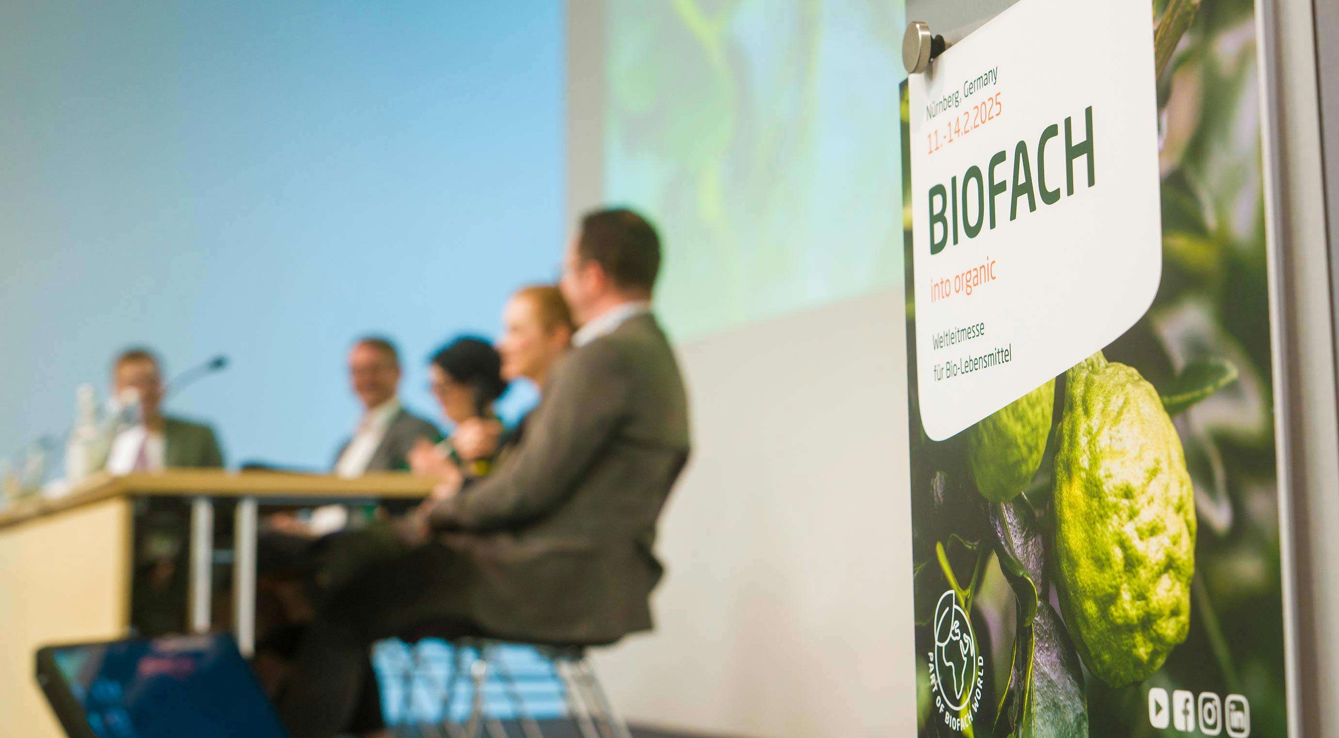 Nahaufnahme eines Schildes der BIOFACH. Im Hintergrund sind unscharf einige Speaker an einem Tisch auf der Bühne zu sehen.